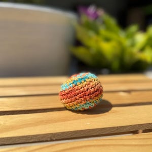 Puede incluir: Una peque&ntilde;a pelota esf&eacute;rica tejida a mano con ganchillo, que presenta un patr&oacute;n de hilo jaspeado con rayas horizontales alternas en naranja brillante, amarillo c&aacute;lido y azul turquesa suave, construida con un punto denso y texturizado.