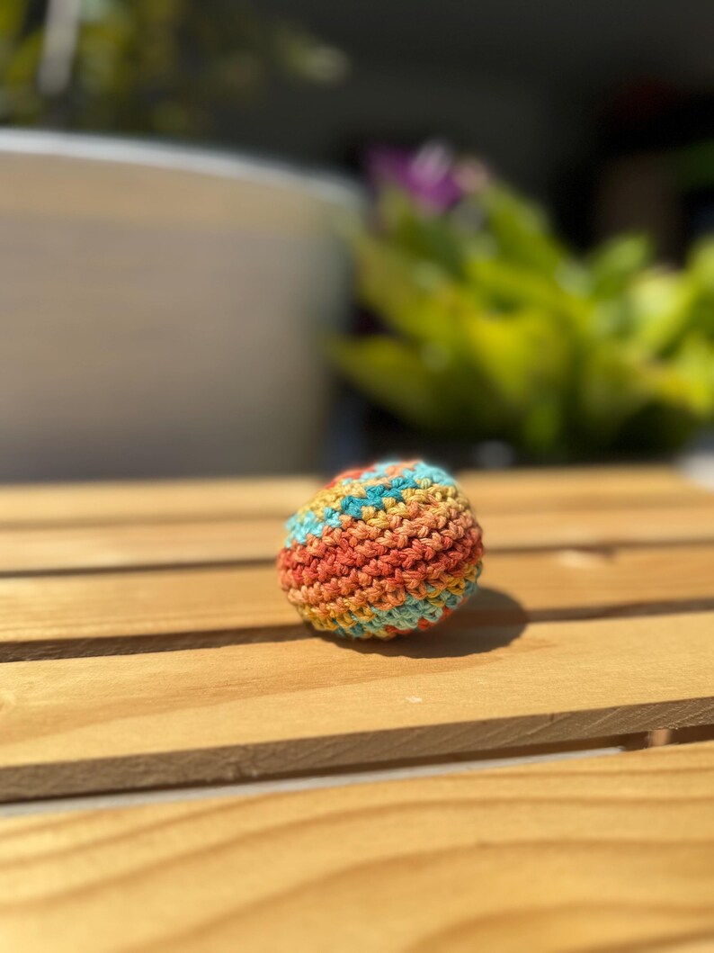 Puede incluir: Una peque&ntilde;a pelota esf&eacute;rica tejida a mano con ganchillo, que presenta un patr&oacute;n de hilo jaspeado con rayas horizontales alternas en naranja brillante, amarillo c&aacute;lido y azul turquesa suave, construida con un punto denso y texturizado.