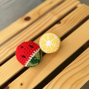 Puede incluir: Dos juguetes de ganchillo con forma de fruta, uno con forma de rodaja de sand&iacute;a con hilo rojo, blanco y verde y detalles de semillas negras, y el otro con forma de rodaja de lim&oacute;n amarillo con hilo blanco que detalla los gajos de la fruta.