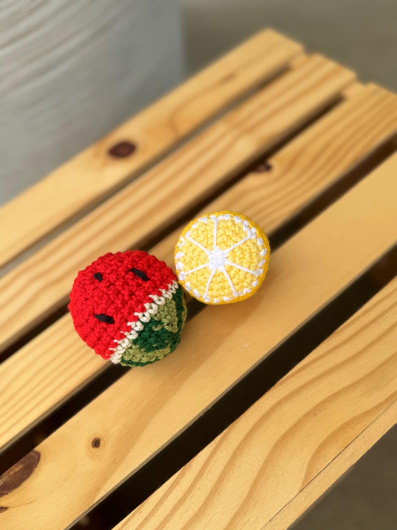 Puede incluir: Dos juguetes de ganchillo con forma de fruta, uno con forma de rodaja de sand&iacute;a con hilo rojo, blanco y verde y detalles de semillas negras, y el otro con forma de rodaja de lim&oacute;n amarillo con hilo blanco que detalla los gajos de la fruta.