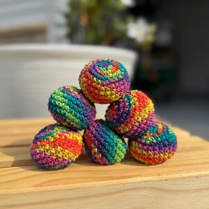 Puede incluir: Una pila de seis peque&ntilde;as bolas esf&eacute;ricas tejidas a ganchillo a mano. Cada bola presenta un patr&oacute;n de hilo matizado vibrante con rayas alternas de rojo brillante, naranja, amarillo, verde, azul y morado, creando una superficie texturizada.