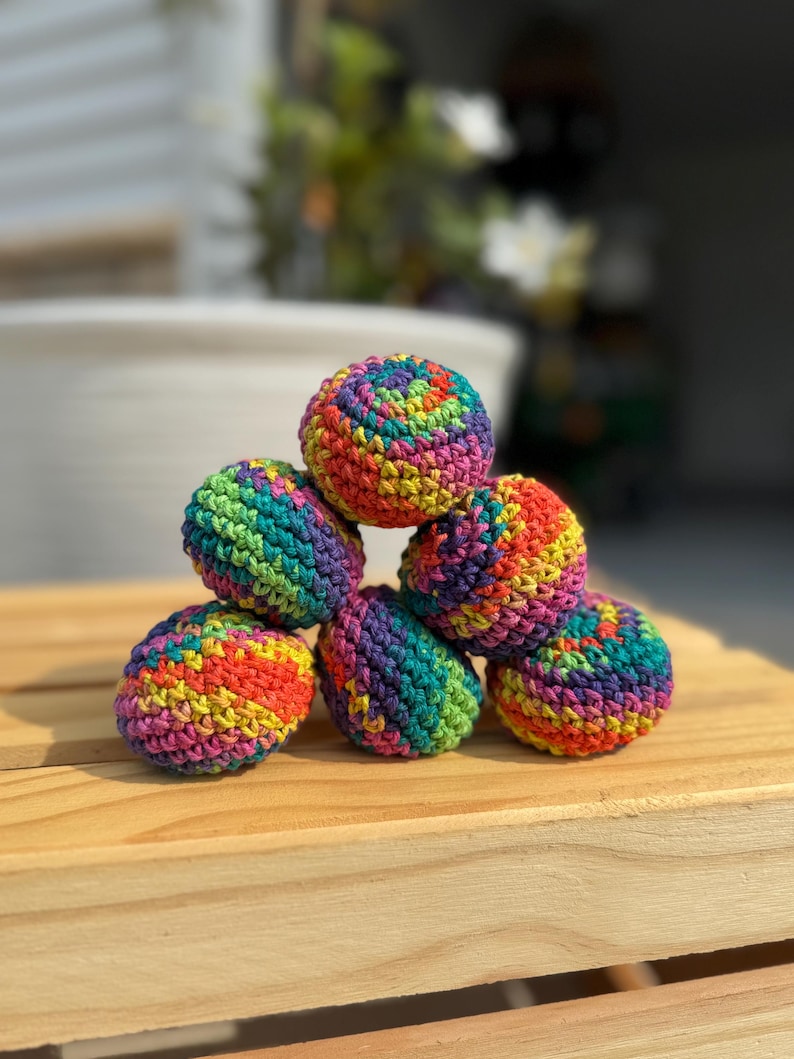 Puede incluir: Una pila de seis peque&ntilde;as bolas esf&eacute;ricas tejidas a ganchillo a mano. Cada bola presenta un patr&oacute;n de hilo matizado vibrante con rayas alternas de rojo brillante, naranja, amarillo, verde, azul y morado, creando una superficie texturizada.