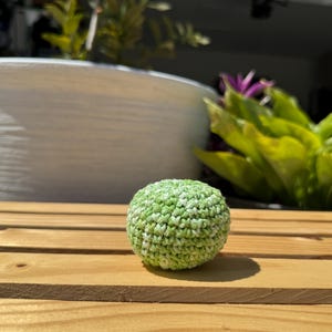 Puede incluir: Un peque&ntilde;o objeto esf&eacute;rico elaborado con hilo verde y blanco jaspeado mediante una t&eacute;cnica de ganchillo. La superficie presenta un patr&oacute;n de puntos texturizado y consistente, creando una forma densa y redondeada con un aspecto moteado.