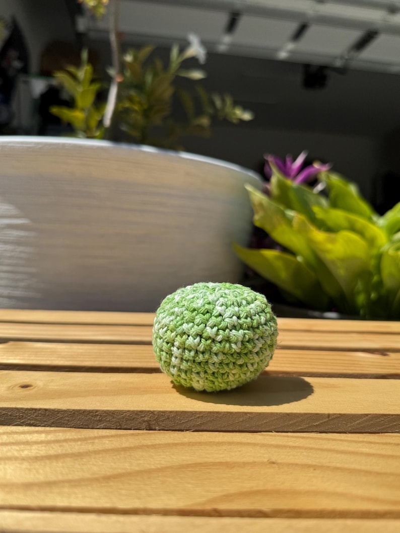 Puede incluir: Un peque&ntilde;o objeto esf&eacute;rico elaborado con hilo verde y blanco jaspeado mediante una t&eacute;cnica de ganchillo. La superficie presenta un patr&oacute;n de puntos texturizado y consistente, creando una forma densa y redondeada con un aspecto moteado.