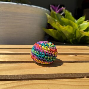 Puede incluir: Un peque&ntilde;o objeto esf&eacute;rico elaborado con hilo multicolor en un patr&oacute;n de punto de ganchillo apretado. La superficie presenta una mezcla vibrante de tonos rojos, naranjas, amarillos, verdes, azules y morados tejidos en una forma densa y redondeada.