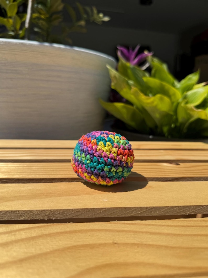 Puede incluir: Un peque&ntilde;o objeto esf&eacute;rico elaborado con hilo multicolor en un patr&oacute;n de punto de ganchillo apretado. La superficie presenta una mezcla vibrante de tonos rojos, naranjas, amarillos, verdes, azules y morados tejidos en una forma densa y redondeada.
