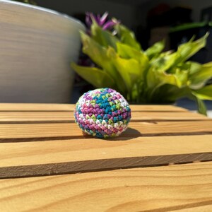 Puede incluir: Un peque&ntilde;o objeto esf&eacute;rico elaborado con hilo multicolor mediante una t&eacute;cnica de ganchillo. La superficie presenta un patr&oacute;n variado de hilos de color verde azulado, verde claro, blanco y morado tejidos estrechamente en una forma redondeada.