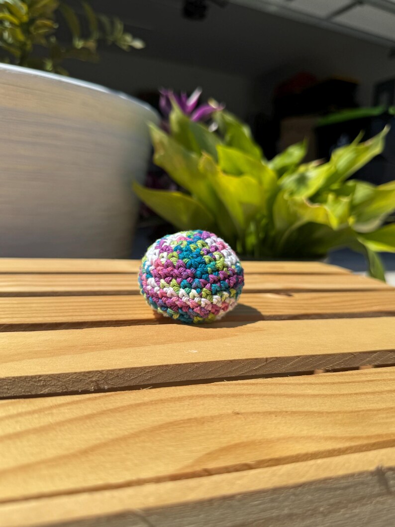 Puede incluir: Un peque&ntilde;o objeto esf&eacute;rico elaborado con hilo multicolor mediante una t&eacute;cnica de ganchillo. La superficie presenta un patr&oacute;n variado de hilos de color verde azulado, verde claro, blanco y morado tejidos estrechamente en una forma redondeada.