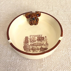 May include: Cream ceramic ashtray with a brown rim and a koala bear figurine. The ashtray has a brown illustration of Sydney landmarks and the city's name.