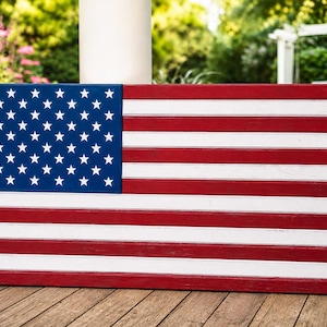 May include: A rectangular wooden wall art piece featuring the design of the American flag. The flag consists of a blue canton with white stars on the left and alternating horizontal red and white stripes extending across the rest of the surface.