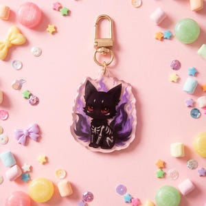 Könnte beinhalten: Ein schwarzer Katzen-Schlüsselanhänger mit Skelett-Design und lila Flammen. Der Schlüsselanhänger hat einen goldenen Verschluss und ist von bunten Süßigkeiten und Dekorationen auf rosa Hintergrund umgeben.