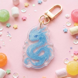 Op de afbeelding: Een heldere acryl sleutelhanger met een lichtblauw drakenontwerp en een goudkleurige sluiting. De sleutelhanger is omgeven door kleurrijke snoepjes, marshmallows, stervormige confetti en pailletten op een roze achtergrond.