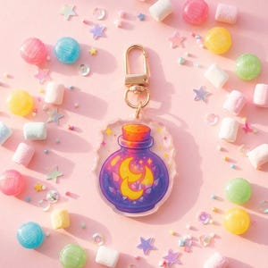 Könnte beinhalten: Ein skurriler Schlüsselanhänger mit einem lila Fläschchen-Design, das einen Mond und Sterne zeigt. Der Schlüsselanhänger hat einen goldfarbenen Verschluss und ist von bunten Süßigkeiten und sternförmigem Konfetti auf rosa Hintergrund umgeben.