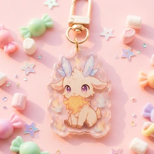 May include: A clear acrylic keychain with a cartoon goat design, crystal horns, and a gold clasp. The goat has a yellow beard and is surrounded by flames. The background is pink with sweets and star-shaped confetti.