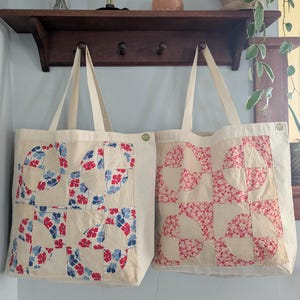 May include: Two cream-colored tote bags with patchwork designs. One features red and blue floral patterns, the other a pink floral pattern. Both bags have long, cream-colored handles and are hanging on a wooden rack.