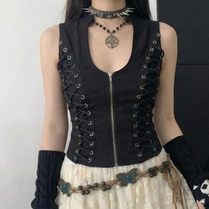 Pode incluir: Top preto sem mangas com atacadores e fecho de correr. O conjunto inclui uma saia de renda branca, um cinto de contas com detalhes de borboletas e luvas sem dedos pretas com desenhos de caveiras. Um colar gargantilha com pontas e um pendente completam o look.