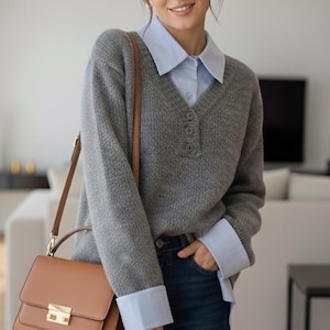 May include: A woman wearing a gray V-neck sweater over a light blue collared shirt, paired with dark blue jeans. A brown leather handbag with a gold clasp hangs from her shoulder. The outfit is completed with gold hoop earrings.