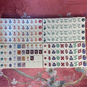 May include: A complete set of ivory-colored Mahjong tiles, arranged in neat rows. The tiles feature various symbols, numbers, and characters in red, blue, green, and black. The set includes tiles with flowers, jokers, and bamboo designs.