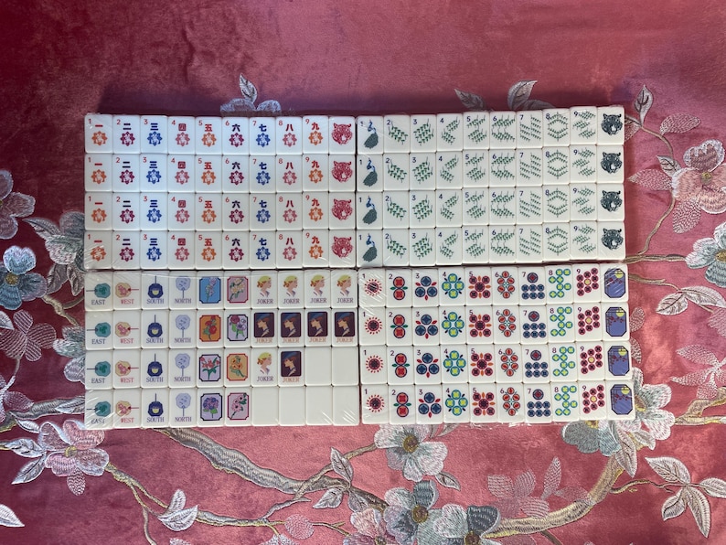 May include: A complete set of ivory-colored Mahjong tiles, arranged in neat rows. The tiles feature various symbols, numbers, and characters in red, blue, green, and black. The set includes tiles with flowers, jokers, and bamboo designs.