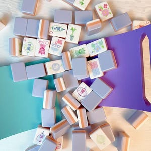 May include: A collection of rectangular mahjong tiles with light purple backs and white faces featuring various colorful illustrations, Chinese characters, and the word Joker, scattered alongside a solid purple plastic tile rack.
