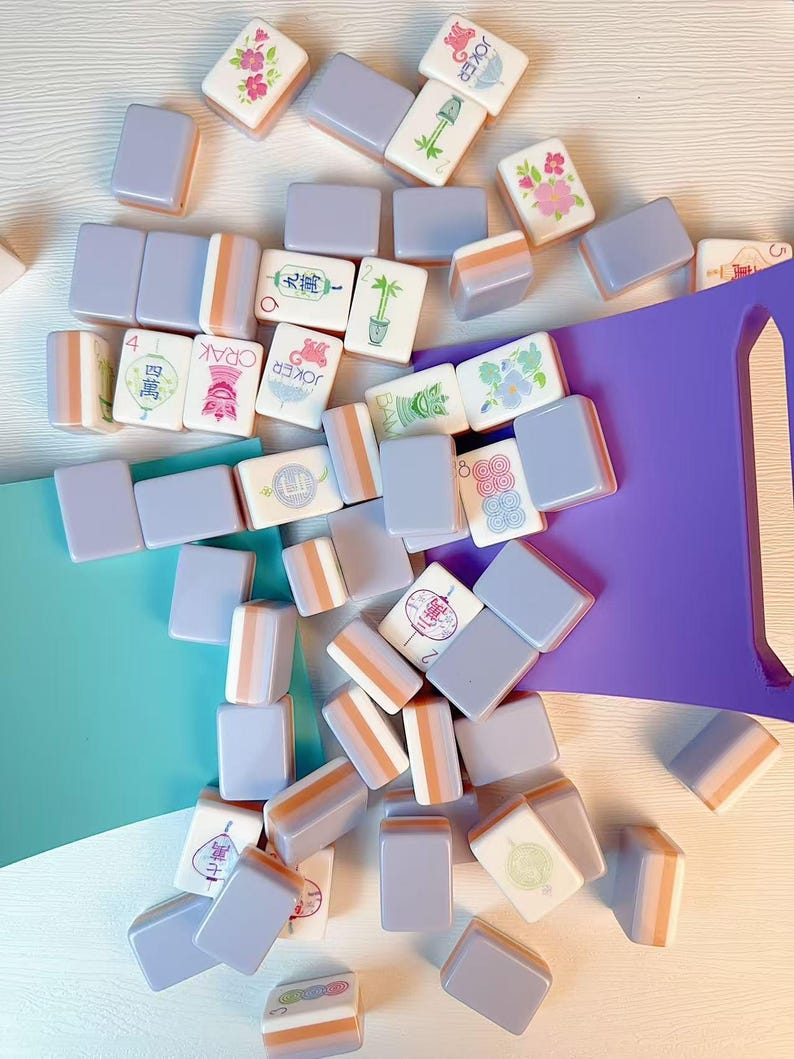 May include: A collection of rectangular mahjong tiles with light purple backs and white faces featuring various colorful illustrations, Chinese characters, and the word Joker, scattered alongside a solid purple plastic tile rack.