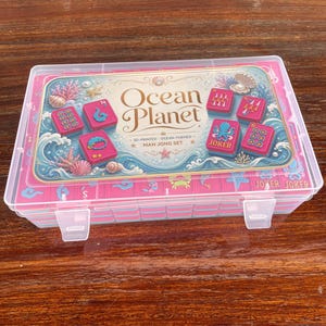 Puede incluir: Una caja de almacenamiento de pl&aacute;stico transparente que contiene un juego de Mah Jong con fichas rosas. La tapa presenta una etiqueta con el texto Ocean Planet, 3D-Printed Ocean-Themed Mah Jong Set e ilustraciones de corales y conchas marinas.
