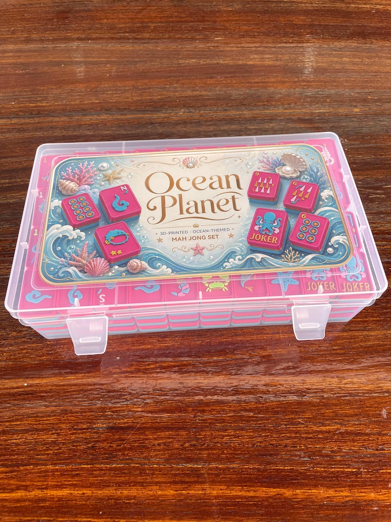 Puede incluir: Una caja de almacenamiento de pl&aacute;stico transparente que contiene un juego de Mah Jong con fichas rosas. La tapa presenta una etiqueta con el texto Ocean Planet, 3D-Printed Ocean-Themed Mah Jong Set e ilustraciones de corales y conchas marinas.