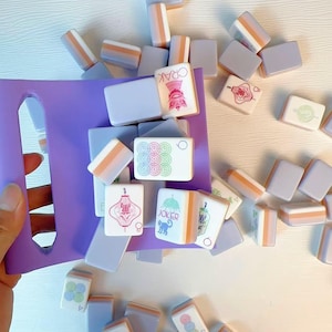 May include: A purple plastic tray holding a collection of rectangular mahjong tiles. The tiles feature light lavender backs with orange accents and white faces displaying various colorful symbols, Chinese characters, and the word JOKER with a cat icon.