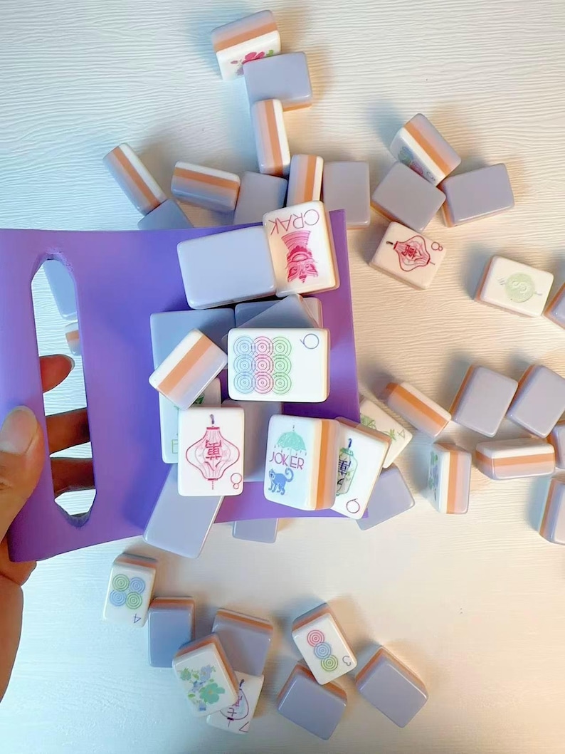 May include: A purple plastic tray holding a collection of rectangular mahjong tiles. The tiles feature light lavender backs with orange accents and white faces displaying various colorful symbols, Chinese characters, and the word JOKER with a cat icon.