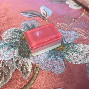 May include: A rectangular Mahjong tile with a translucent pink top, a light pink middle layer, and a white base. The tile rests on a pink floral embroidered fabric. The tile is part of a Mahjong set.