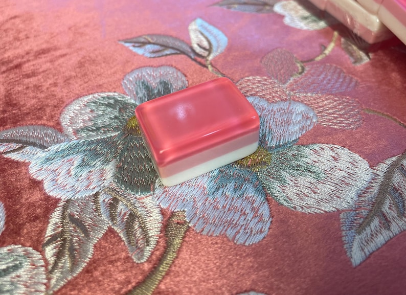 May include: A rectangular Mahjong tile with a translucent pink top, a light pink middle layer, and a white base. The tile rests on a pink floral embroidered fabric. The tile is part of a Mahjong set.