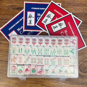 May include: A clear plastic case containing a set of Mahjong tiles with Christmas-themed illustrations, including ornaments, stockings, and gifts. Behind the case are several red and blue cards labeled Standard Hands and Rules 2026.