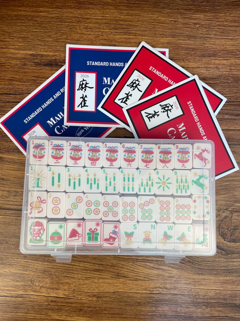 May include: A clear plastic case containing a set of Mahjong tiles with Christmas-themed illustrations, including ornaments, stockings, and gifts. Behind the case are several red and blue cards labeled Standard Hands and Rules 2026.