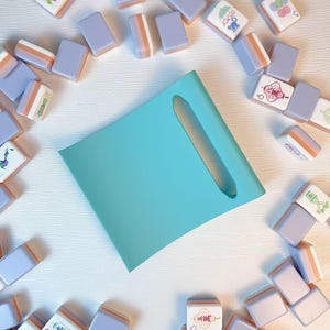 May include: A light turquoise rectangular tray with a central elongated cutout, surrounded by a scattered collection of mahjong tiles. The tiles feature a lavender back and white face with various colorful illustrations and Chinese characters.