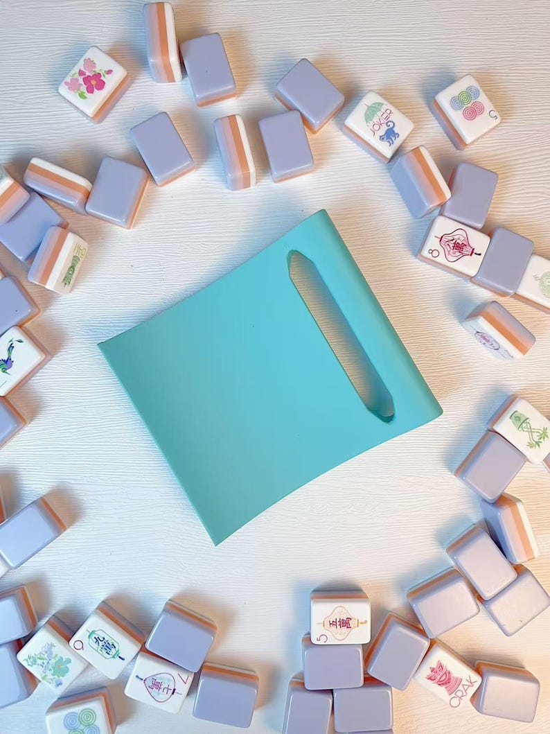 May include: A light turquoise rectangular tray with a central elongated cutout, surrounded by a scattered collection of mahjong tiles. The tiles feature a lavender back and white face with various colorful illustrations and Chinese characters.