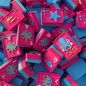 Puede incluir: Una colecci&oacute;n de fichas de juego rectangulares con una superficie superior rosa brillante y una base azul claro. Las fichas muestran varios dise&ntilde;os en relieve en azul, amarillo y blanco, incluyendo una estrella de mar, un pulpo con la etiqueta JOKER y n&uacute;meros.