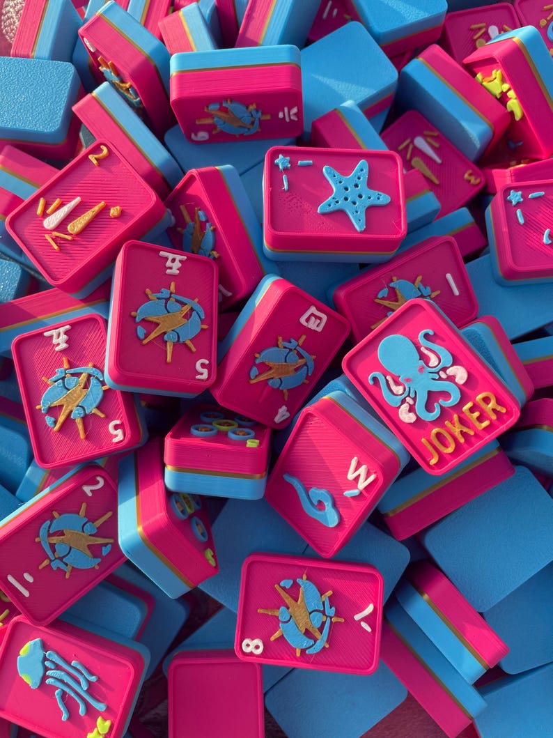 Puede incluir: Una colecci&oacute;n de fichas de juego rectangulares con una superficie superior rosa brillante y una base azul claro. Las fichas muestran varios dise&ntilde;os en relieve en azul, amarillo y blanco, incluyendo una estrella de mar, un pulpo con la etiqueta JOKER y n&uacute;meros.