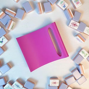 May include: A bright pink rectangular tile rack with a vertical slot, surrounded by a collection of scattered mahjong tiles in shades of lavender and white, featuring various colorful illustrations and characters on their faces.