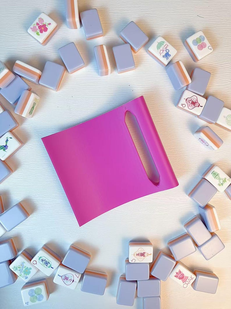 May include: A bright pink rectangular tile rack with a vertical slot, surrounded by a collection of scattered mahjong tiles in shades of lavender and white, featuring various colorful illustrations and characters on their faces.