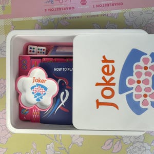 May include: A white plastic box containing a board game. The box is open, revealing dice, game cards, and a flower-shaped dish with the word "Joker" on it. The lid features the word "Joker" in orange and a blue and pink floral design.