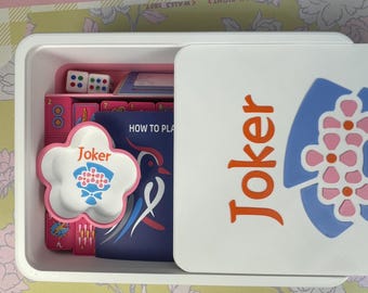Könnte beinhalten: Eine weiße Plastikbox mit einem Brettspiel. Die Box ist geöffnet und zeigt Würfel, Spielkarten und eine blütenförmige Schale mit dem Wort "Joker". Der Deckel zeigt das Wort "Joker" in Orange und ein blaues und rosa Blumenmuster.