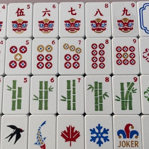 Op de afbeelding: Een verzameling witte rechthoekige mahjongtegels met diverse ontwerpen, waaronder Chinese cijfers, rode leeuwenkoppen, cirkelvormige muntpatronen, groene bamboestengels, een zwarte vogel, een koikarper, een rood esdoornblad, een blauwe sneeuwvlok en een joker.