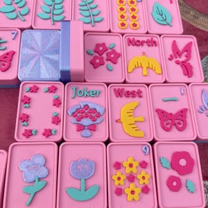 May include: A set of pink Mahjong tiles with various colorful designs. The tiles feature floral, leaf, and bird motifs, along with the words "Joker" and "West". The set includes a pink and blue tile holder.