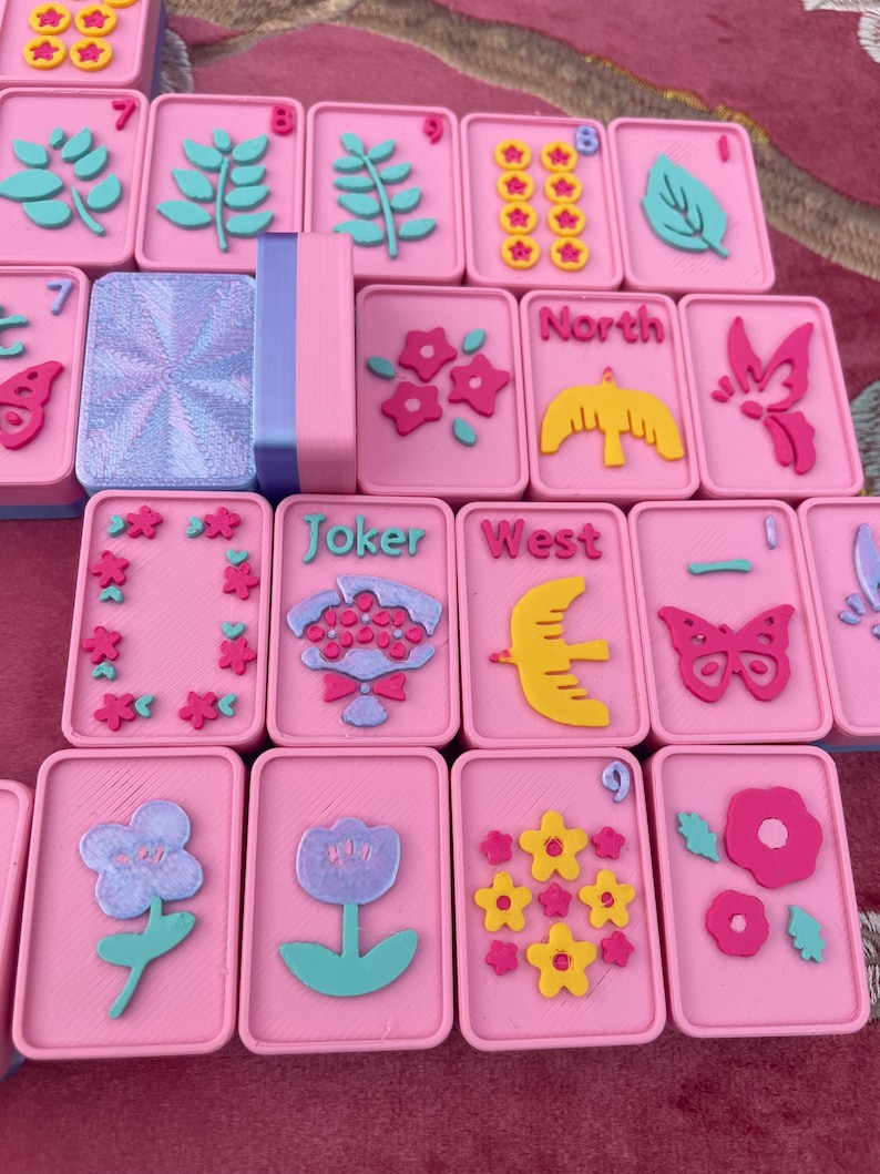 May include: A set of pink Mahjong tiles with various colorful designs. The tiles feature floral, leaf, and bird motifs, along with the words "Joker" and "West". The set includes a pink and blue tile holder.