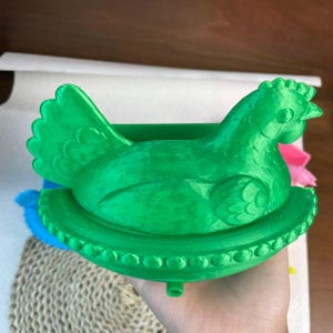 May include: A bright green chicken-shaped container, possibly a decorative item or storage solution. The chicken is perched on a round base with a beaded edge. The container is held in a hand.