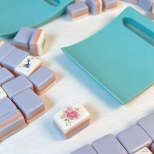 May include: A set of rectangular mahjong tiles featuring a lavender and peach color scheme with white faces. Some tiles display floral and character illustrations. Two teal plastic tile racks are positioned among the scattered game pieces.