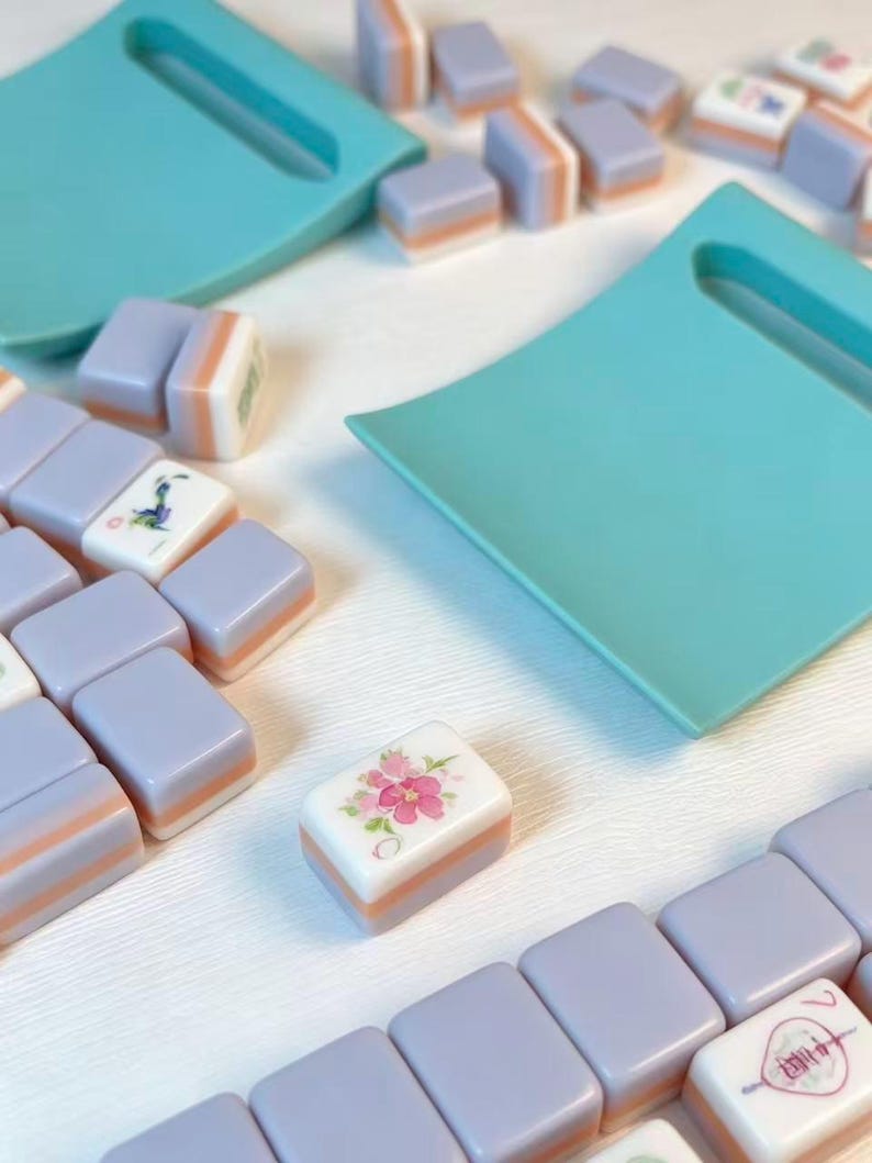 May include: A set of rectangular mahjong tiles featuring a lavender and peach color scheme with white faces. Some tiles display floral and character illustrations. Two teal plastic tile racks are positioned among the scattered game pieces.