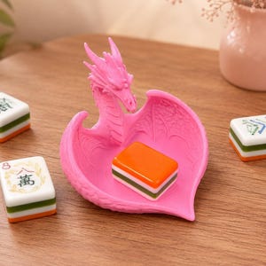 Pu&ograve; includere: Un piattino portagioie rosa brillante a forma di drago, con testa e ali scolpite che formano una coppa. Una tessera rettangolare del mahjong arancione, bianca e verde riposa all'interno, mentre altre due tessere si trovano nelle vicinanze.