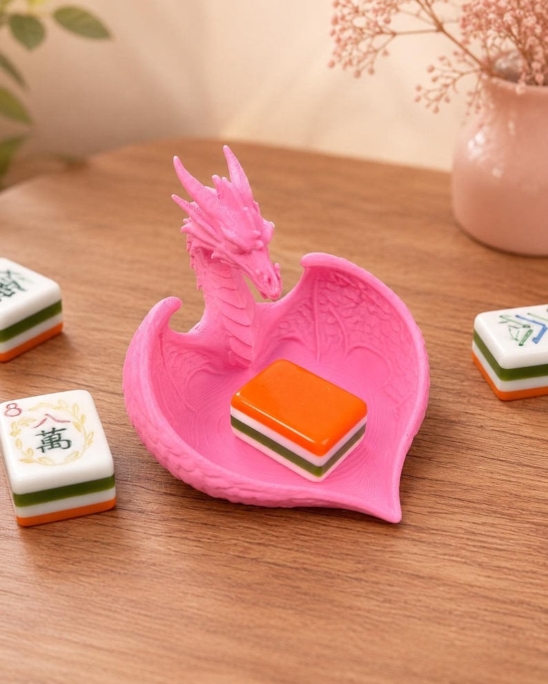 Pu&ograve; includere: Un piattino portagioie rosa brillante a forma di drago, con testa e ali scolpite che formano una coppa. Una tessera rettangolare del mahjong arancione, bianca e verde riposa all'interno, mentre altre due tessere si trovano nelle vicinanze.