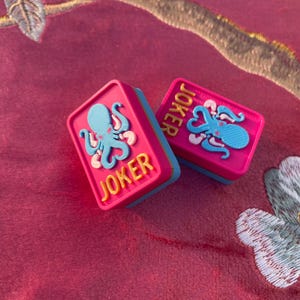 Puede incluir: Dos fichas de juego rectangulares con una superficie superior de color rosa brillante, una base azul claro y un gr&aacute;fico de pulpo azul claro en relieve. La palabra JOKER est&aacute; grabada en letras amarillas sobre la superficie rosa de cada ficha.